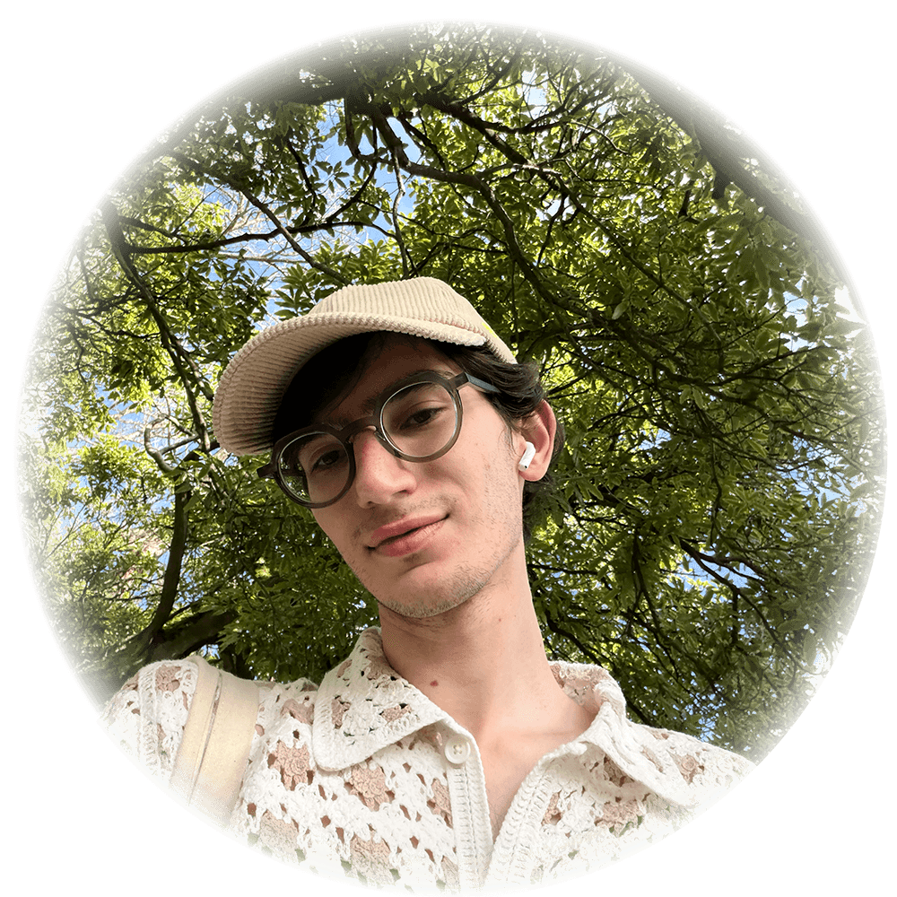 Person with round glasses wearing a lace shirt and a corduroy cap with verdant foliage behind them.