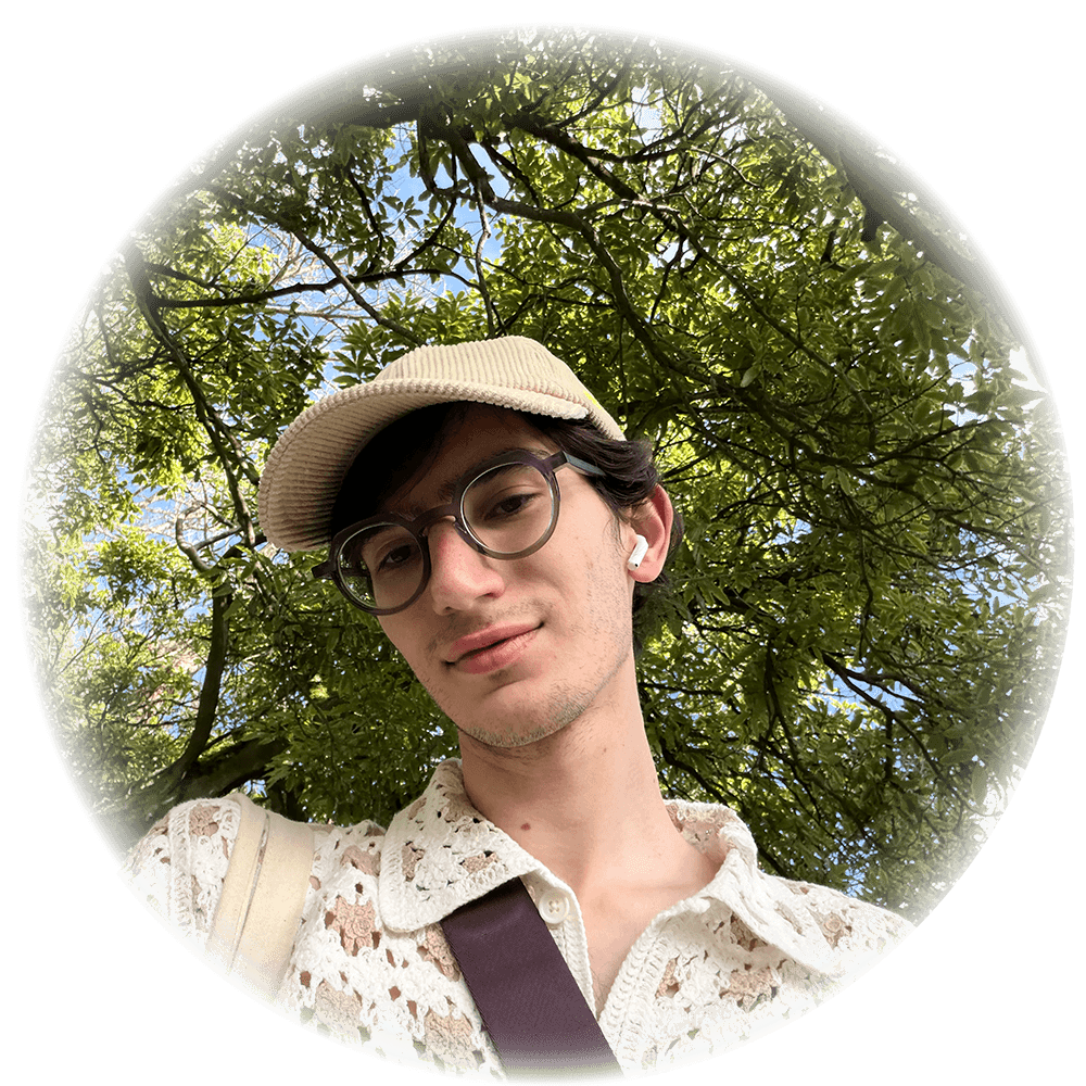 Person with round glasses wearing a lace shirt and a corduroy cap with verdant foliage behind them.