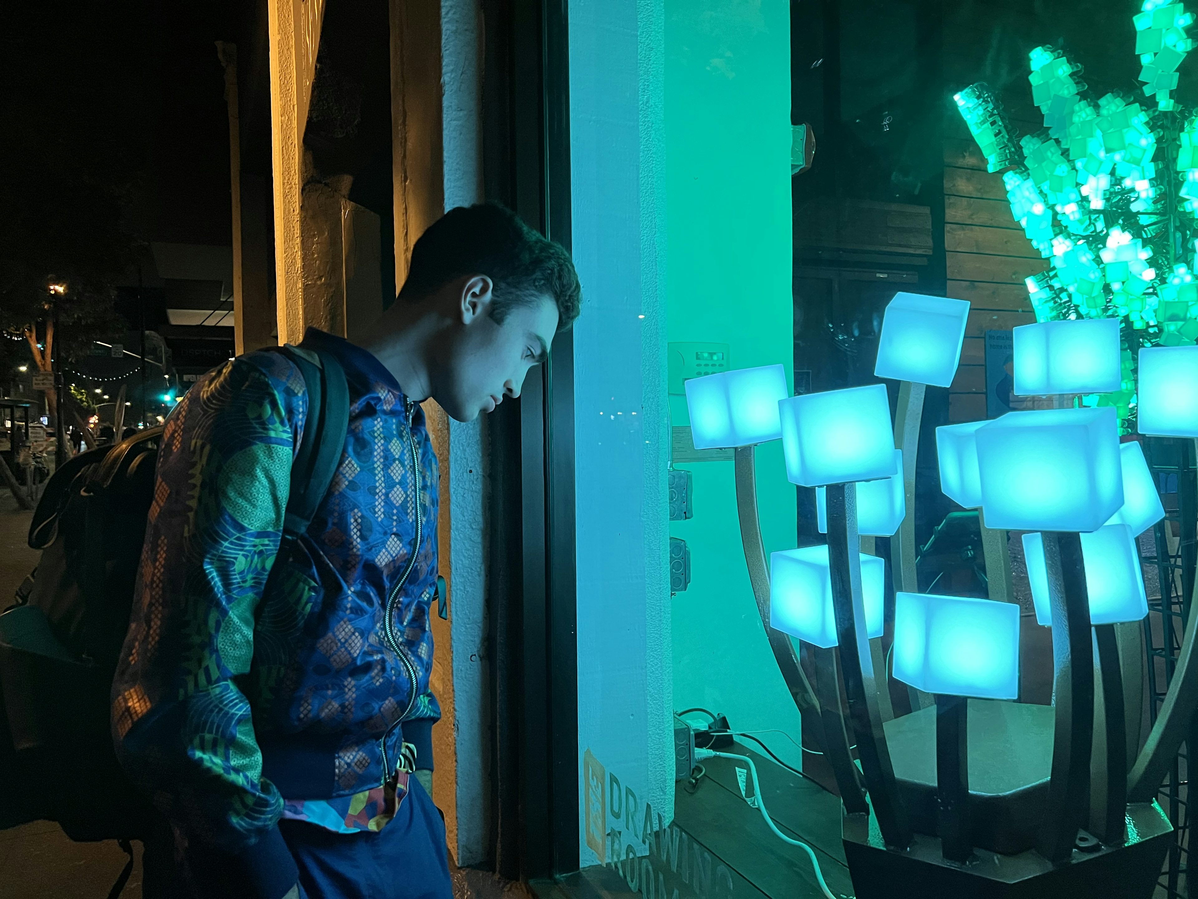 Lachlan wearing a reflective bomber jacket appearing moody looking at an art exhibit in a window with green lighting
